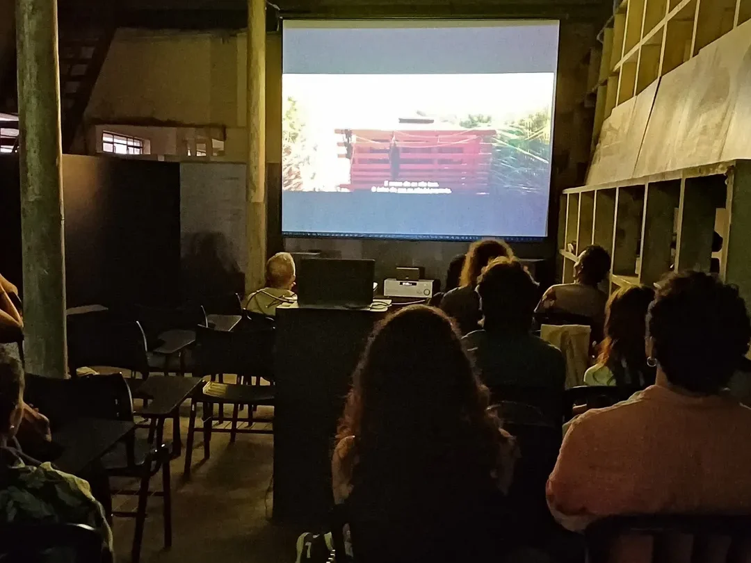 Pessoas sentadas em cadeiras assistem a um filme projetado em uma tela grande em um ambiente simples e escuro.