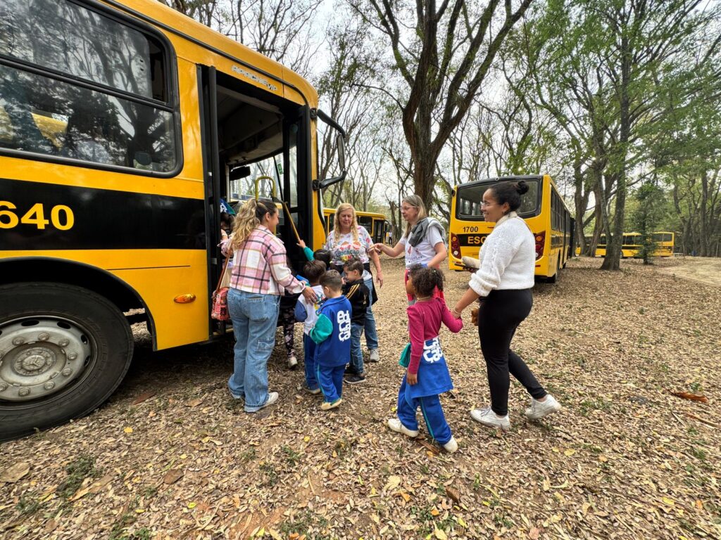 Crianças pequenas, acompanhadas de adultos, descem de um ônibus escolar amarelo em uma área arborizada. Ao fundo, outros ônibus estacionados aguardam.