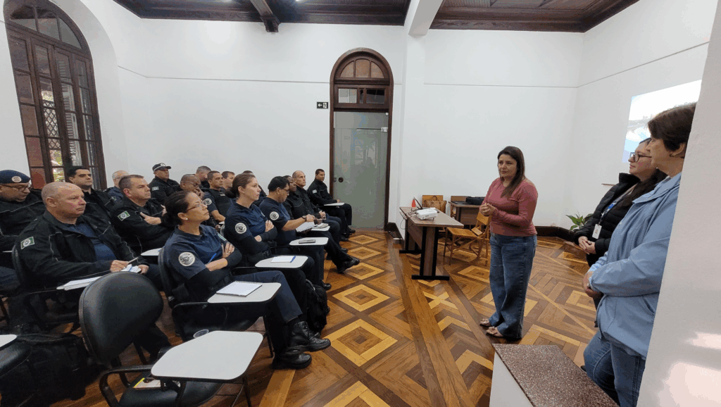 A imagem mostra uma sala de aula com um grupo de pessoas sentadas em cadeiras, a maioria vestindo uniformes escuros. Há uma mulher em pé na frente da sala, falando com o grupo. Ela está vestindo uma camisa rosa e calças jeans. Atrás dela, há um projetor e uma tela com uma imagem. A sala tem piso de madeira com um padrão geométrico e paredes brancas. Há uma grande janela com molduras escuras em um lado da sala e uma porta com uma moldura de madeira semelhante no outro lado. A atmosfera parece ser de uma palestra ou treinamento.