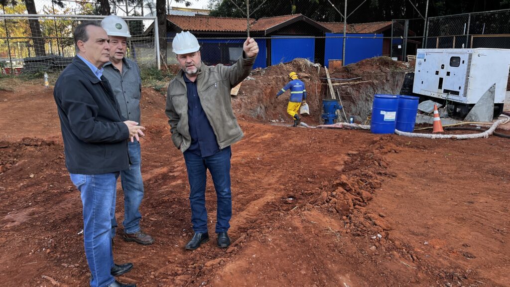 Na imagem, três homens em um canteiro de obras. Dois deles, à esquerda, usam capacetes brancos e estão conversando. O homem da frente usa um casaco preto sobre uma camisa azul e calça jeans, com sapatos pretos. O homem atrás dele usa uma camisa xadrez e calça jeans, com sapatos marrons. O terceiro homem, no centro, também usa um capacete branco e um casaco marrom sobre uma camisa azul escura e calça jeans, com sapatos pretos. Ele está gesticulando com a mão direita, como se estivesse explicando algo. Ao fundo, um trabalhador com capacete amarelo e vestimenta de segurança amarela e azul está próximo a uma escavação. Há também equipamentos como um gerador branco, dois barris azuis e mangueiras. O terreno é coberto por terra vermelha, indicando uma área de construção.