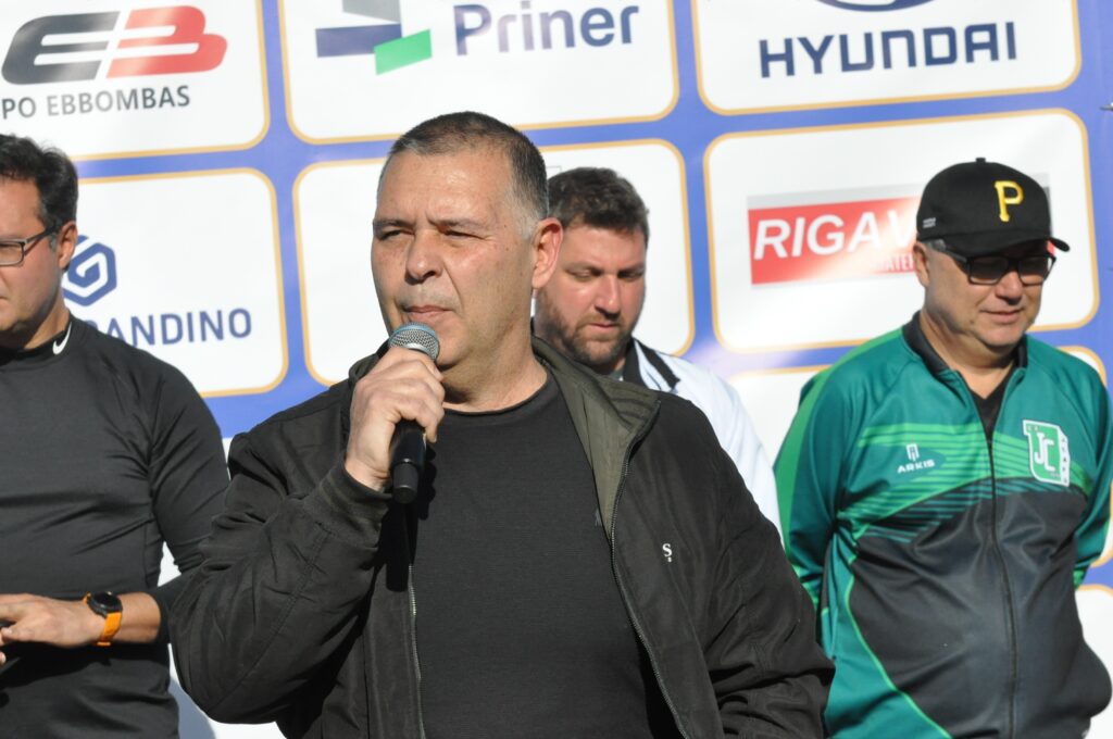A imagem mostra um grupo de homens em pé em frente a um painel com logotipos de patrocinadores. O homem em primeiro plano, no centro da imagem, está segurando um microfone e falando. Ele tem cabelo curto e escuro e veste uma jaqueta preta sobre uma camiseta preta. À sua esquerda, um homem com óculos e cabelo escuro veste uma camiseta preta com um logotipo no peito. À direita do homem que fala, há outro homem com barba e cabelo escuro, vestindo uma camisa branca com detalhes em preto. Mais à direita, um homem com óculos e um boné preto com a letra "P" amarela está vestindo uma jaqueta verde e preta com um logotipo no peito. O painel atrás deles exibe logotipos de empresas como "EB", "Priner", "HYUNDAI" e "RIGAMONTI". A iluminação sugere que a foto foi tirada ao ar livre durante o dia.