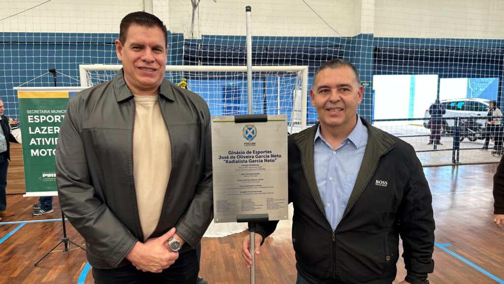 A imagem mostra dois homens em pé em uma quadra esportiva interna, provavelmente um ginásio. Eles estão posando em frente a uma placa comemorativa que está fixada em um pedestal.À esquerda, um homem veste uma jaqueta de couro preta sobre uma camiseta bege e calças escuras. Ele usa um relógio no pulso esquerdo e tem as mãos cruzadas na frente do corpo. Ele sorri para a câmera.À direita, outro homem está em pé, vestindo uma jaqueta escura sobre uma camisa azul de botões. Ele também sorri para a câmera. A jaqueta dele tem o logotipo "BOSS" no peito.A placa em destaque tem o brasão da cidade de Piracicaba no topo e o seguinte texto em português: "Ginásio de Esportes José de Oliveira Garcia Neto 'Radialista Garcia Neto'". Abaixo, há detalhes sobre a reforma do ginásio, incluindo a data de entrega e nomes de pessoas envolvidas, como o prefeito e secretários.