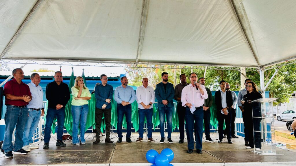 A imagem mostra um grupo de pessoas em pé em um palco montado sob uma tenda branca. A maioria das pessoas está vestida formalmente, com camisas e calças. Há algumas bandeiras verdes ao fundo, e árvores e céu visíveis acima da tenda. Uma mulher está em pé atrás de um púlpito transparente, segurando um copo e um pedaço de papel, enquanto um homem à sua frente fala em um microfone. O homem que fala está vestindo uma camisa rosa clara e calça azul. Há também três balões azuis no chão, em primeiro plano.