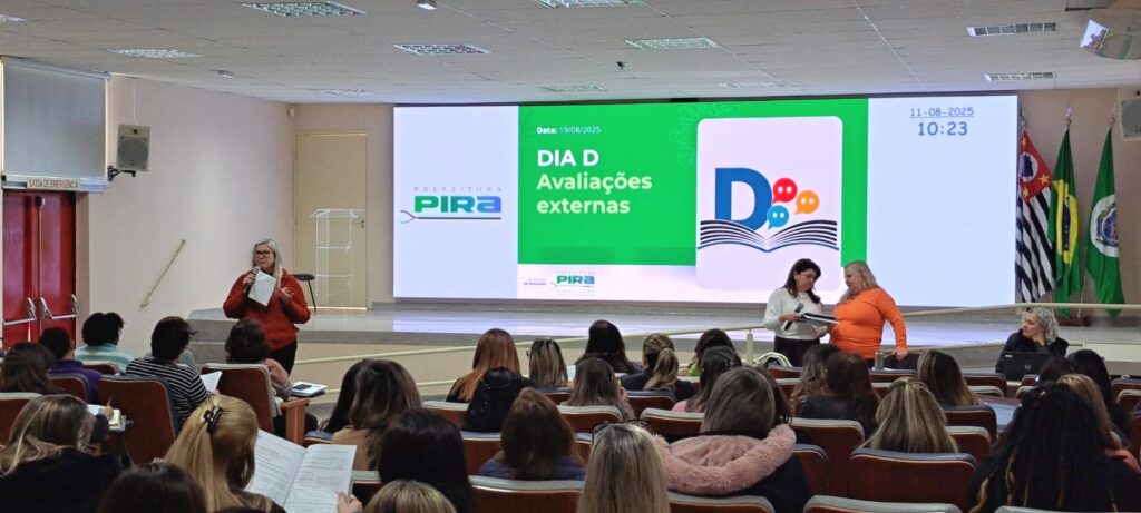A imagem retrata uma sala de conferências onde ocorre um evento intitulado “DIA D Avaliações externas”, conforme indicado no grande telão ao fundo, que também exibe a data “19/08/2025” e o logotipo da Prefeitura de Pira. O ambiente está organizado com cadeiras em fileiras, ocupadas por participantes atentos, muitos com documentos ou cadernos em mãos. Duas pessoas estão em pé diante do público — uma com microfone e outra com papéis — conduzindo a atividade. Ao lado do telão, destacam-se as bandeiras do Brasil, do estado de São Paulo e de um município, reforçando o caráter institucional do encontro.