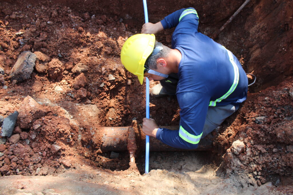 Um trabalhador com capacete amarelo e uniforme azul com detalhes fluorescentes está agachado em uma vala. Ele está segurando uma haste azul e examinando um cano enferrujado que está parcialmente exposto na terra. A terra ao redor é marrom e pedregosa, com alguns pedaços de concreto. O sol está brilhando, criando sombras fortes.