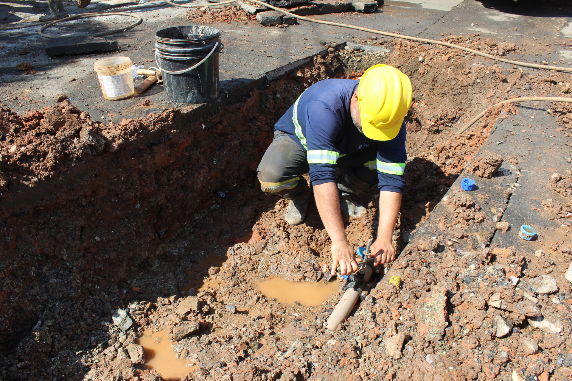 Um trabalhador com capacete amarelo e uniforme azul está agachado em uma vala de terra, realizando reparos em um cano. O cano, que parece ser de PVC, está parcialmente submerso em água lamacenta. O trabalhador está usando luvas e segurando uma ferramenta para apertar uma conexão no cano. Ao fundo, há um balde preto e um recipiente de plástico aberto, possivelmente contendo ferramentas ou materiais. A área ao redor está coberta de terra solta, pedras e pedaços de asfalto. Fios e mangueiras também são visíveis na superfície. O sol está forte, criando sombras distintas na cena.