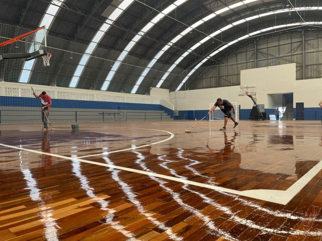 A imagem mostra o interior de uma quadra poliesportiva com piso de madeira brilhante. Dois homens estão na quadra, cada um com um rodo, limpando ou aplicando algum produto no piso. O teto da quadra é abobadado, com diversas aberturas para iluminação natural. Há cestas de basquete em ambas as extremidades da quadra e redes de proteção nas laterais. O piso está molhado e reflete a luz, criando um efeito espelhado.