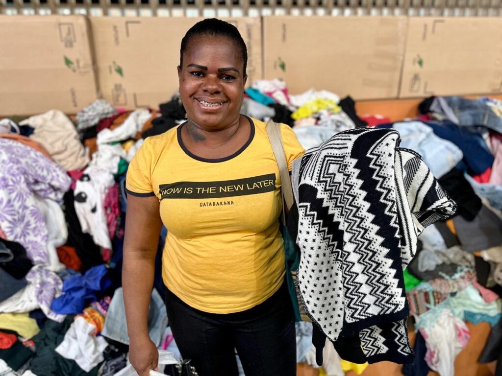 A mulher sorri para a câmera, segurando um suéter preto e branco com um padrão geométrico. Ela está vestindo uma camiseta amarela com a frase "NOW IS THE NEW LATER" e "GATABAKANA" escrita nela. Ao fundo, há uma grande pilha de roupas.