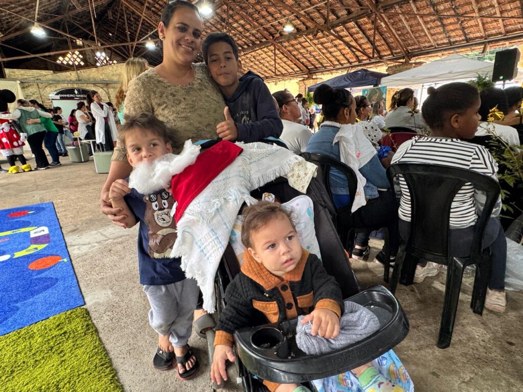 A imagem mostra um grupo de pessoas reunidas em um evento ao ar livre, possivelmente uma feira ou festival. No primeiro plano, uma mulher sorri enquanto abraça dois meninos. Um dos meninos, mais velho, está com o polegar para cima, indicando aprovação. O outro menino, mais novo, está no colo da mulher e parece estar vestindo uma fantasia, com uma barba branca e um chapéu vermelho, possivelmente de Papai Noel. Ele está segurando um cobertor ou manta. Ao lado deles, em um carrinho de bebê, um bebê com um casaco preto e marrom está sentado, olhando para o lado. O carrinho possui uma bandeja com um cobertor cinza. Ao fundo, mais pessoas estão sentadas em cadeiras, assistindo ao evento. Algumas figuras fantasiadas, como o Mickey Mouse, estão visíveis à esquerda, interagindo com o público. Há também tendas e uma estrutura de madeira com telhado aparente, sugerindo um ambiente de evento. A atmosfera parece ser festiva e familiar.