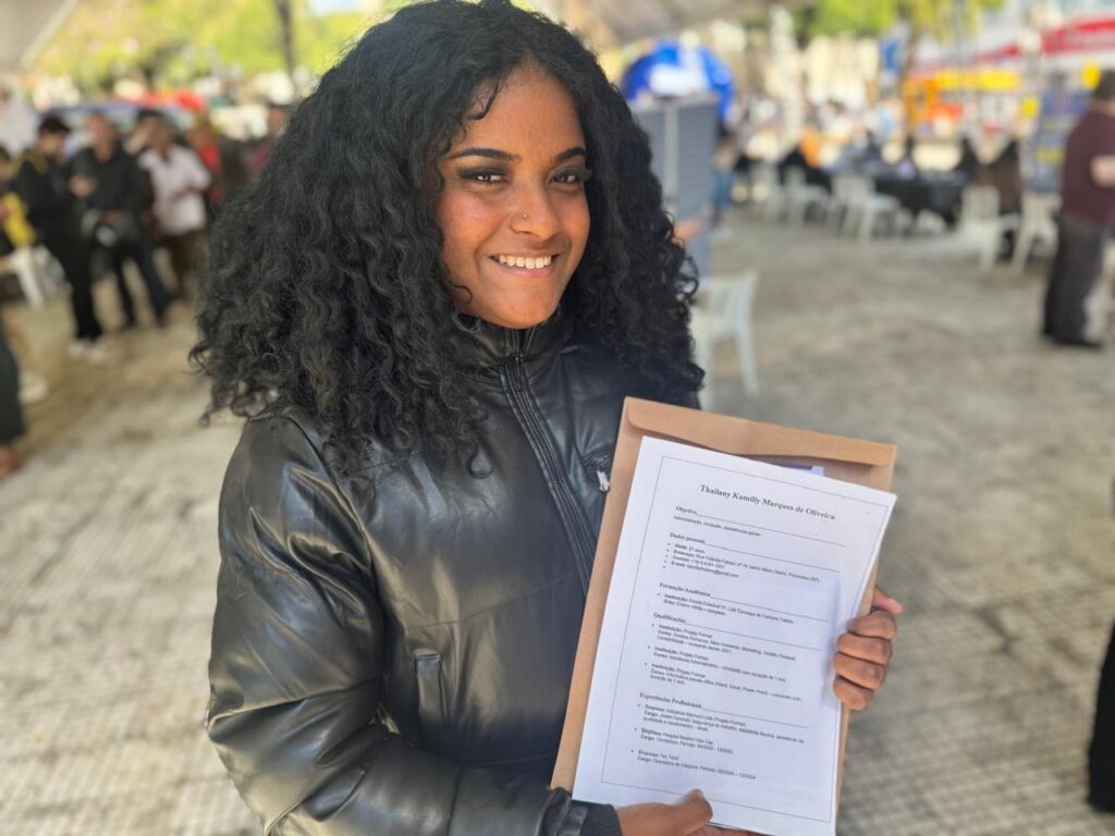 A imagem mostra uma jovem mulher sorrindo para a câmera, segurando um currículo em um envelope. Ela tem cabelos escuros e cacheados e está usando uma jaqueta preta. O fundo está desfocado, mas parece ser um evento ao ar livre com outras pessoas e tendas. O currículo que ela está segurando tem o nome "Thailany Kamilly Marques de Oliveira" no topo.