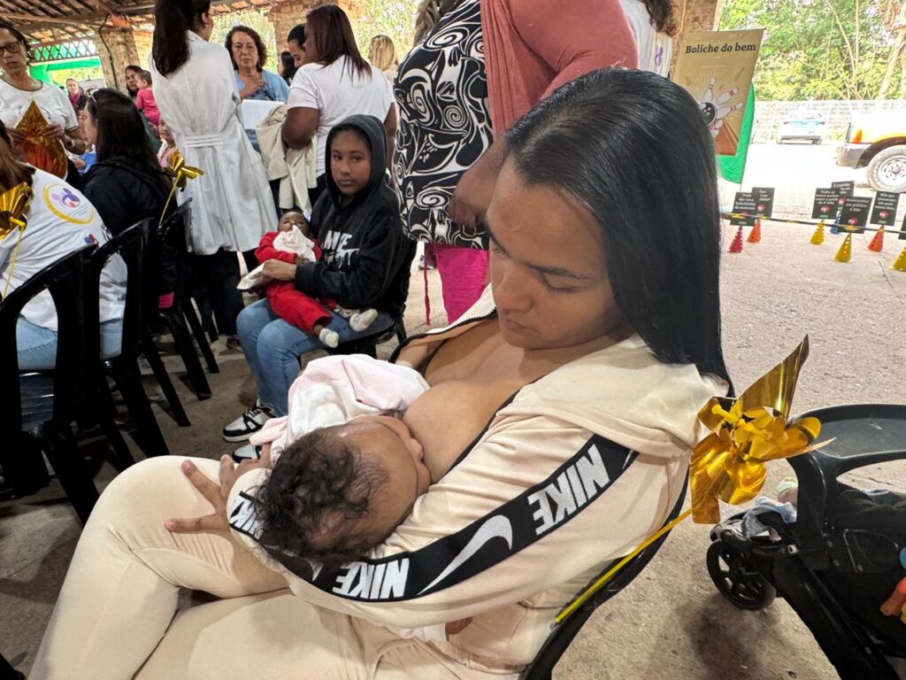 A imagem mostra um grupo de mulheres reunidas em um evento ao ar livre. No primeiro plano, uma mulher está amamentando um bebê. Ela está usando um agasalho bege com a marca "NIKE" em destaque. O bebê, com cabelo escuro, está aconchegado em seus braços. Ao fundo, outras mulheres estão sentadas em cadeiras, observando o evento. Algumas delas usam camisetas brancas com um símbolo de amamentação, sugerindo que o evento pode estar relacionado à amamentação ou ao apoio à maternidade. Há também uma menina sentada em uma cadeira, segurando um bebê vestido de vermelho. O ambiente parece ser um local com cobertura de telhado, possivelmente um pátio ou área de eventos. Há uma placa com a inscrição "Boliche do bem" e imagens de boliche, indicando que o evento pode ter alguma conexão com essa atividade ou ser um evento de arrecadação de fundos. Pequenos cones coloridos e placas com textos estão dispostos no chão, adicionando um toque festivo ao local. Uma cadeira de rodas ou carrinho de bebê está parcialmente visível à direita.