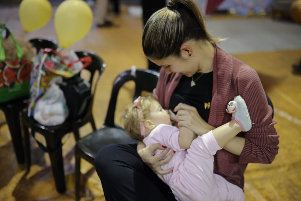 A mulher está sentada em uma cadeira preta, com um bebê em seu colo. Ela está amamentando o bebê, que está vestido com uma roupa rosa. A mulher está usando um cardigã rosa por cima de uma blusa preta. Ela tem o cabelo preso em um rabo de cavalo. Há dois balões amarelos ao fundo, e algumas sacolas em outras cadeiras.