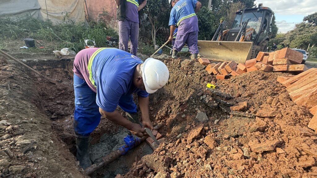 A imagem mostra uma cena de trabalho em uma vala, com vários trabalhadores envolvidos em uma tarefa de reparo ou instalação de tubulação subterrânea. No primeiro plano, um trabalhador está ajoelhado dentro da vala, usando um capacete branco e um uniforme azul com detalhes em verde e amarelo. Ele está concentrado em uma tubulação de água azul com uma válvula, utilizando uma ferramenta manual para trabalhar em uma seção da tubulação, possivelmente para reparo ou conexão. A vala está preenchida com terra solta e escombros, incluindo pedras e pedaços de asfalto. Ao fundo, outros dois trabalhadores estão visíveis. Um deles, com um uniforme semelhante, está mais atrás e parece estar manuseando uma pá ou ferramenta semelhante. O outro trabalhador, também de capacete e uniforme azul, está em pé e parece estar supervisionando ou auxiliando na tarefa. Um trator carregador (retroescavadeira) está posicionado ao fundo, com a pá levantada, sugerindo que o maquinário está sendo usado para mover terra ou materiais no local. Há uma pilha de tijolos vermelhos dispostos no chão, perto da borda da vala, indicando que eles podem ser usados ​​na obra. Alguns pedaços de asfalto e terra também estão espalhados pela área. O ambiente parece ser um local externo, possivelmente uma rua ou área de serviço, com vegetação e uma construção ao fundo. O céu está visível, com algumas nuvens. A iluminação sugere que é dia.