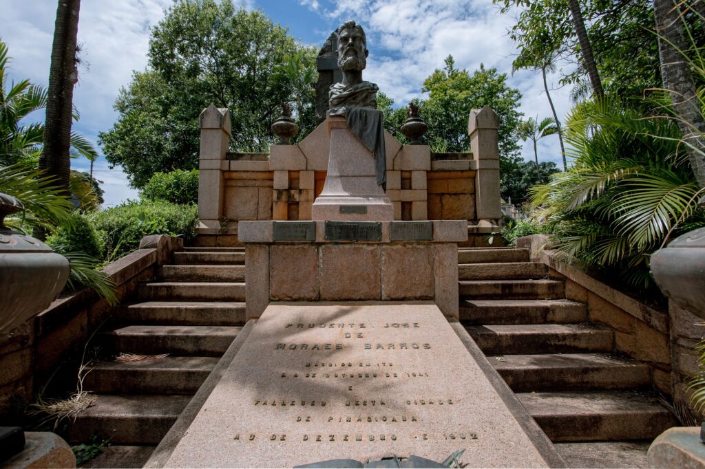 A imagem mostra um monumento com uma escadaria de pedra que leva a um busto de um homem barbudo. O monumento é feito de pedra e tem várias placas com inscrições.A escadaria é larga e íngreme, com corrimãos de pedra em ambos os lados. Há vasos de pedra em cada lado da base da escadaria.O busto está colocado sobre um pedestal de pedra, que por sua vez está sobre uma estrutura maior de pedra. Atrás do busto, há uma parede de pedra com nichos em ambos os lados, cada um contendo um vaso decorativo.A inscrição principal na base do monumento, na parte inferior da imagem, diz:PRUDENTE JOSE
DE
MORAES BARROSNASCEU EM 1834
E FALECEU NESTA CIDADE
DE PIRACICABA
A 9 DE DEZEMBRO DE 1902O monumento está cercado por vegetação exuberante, incluindo árvores altas e palmeiras. O céu está parcialmente nublado.