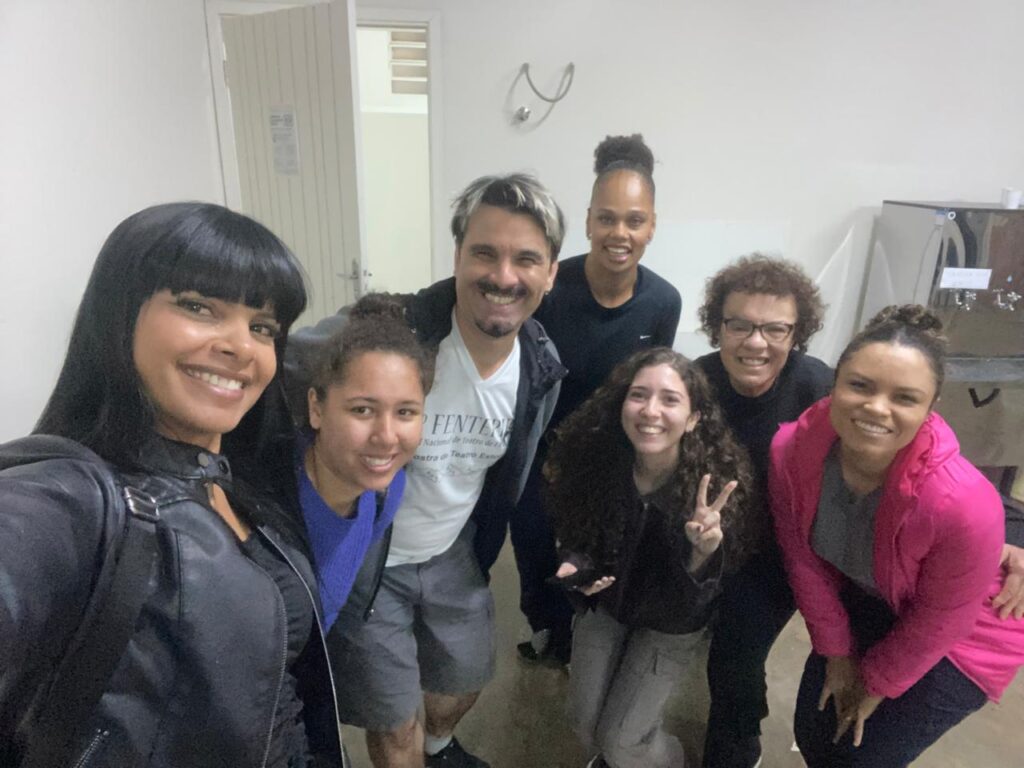 Grupo de sete pessoas posa sorrindo para uma selfie em ambiente interno, com expressão de alegria.