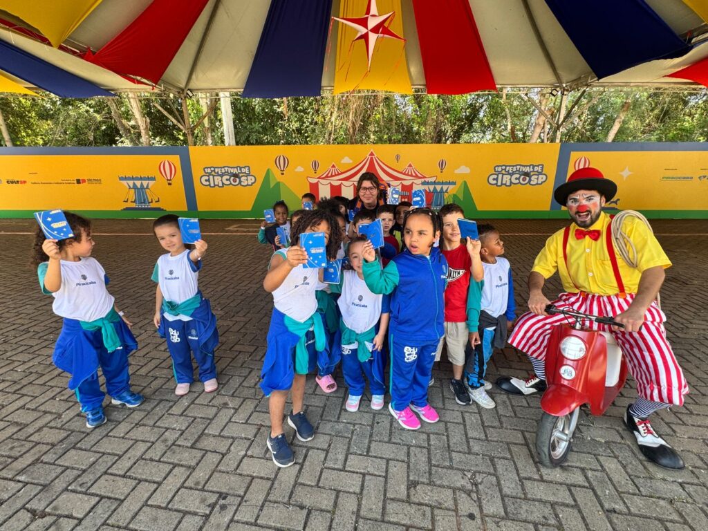 Pra cego ver: Crianças pequenas, do Jardim I da Escola Municipal Enedina Lourenço Vieira, posam sorridentes ao lado de um palhaço. Elas usam uniforme azul e branco da Prefeitura de Piracicaba e seguram caderninhos azuis do Festival de Circo. O palhaço veste calça listrada vermelha e branca, camiseta amarela, suspensório vermelho e gravata borboleta vermelha.