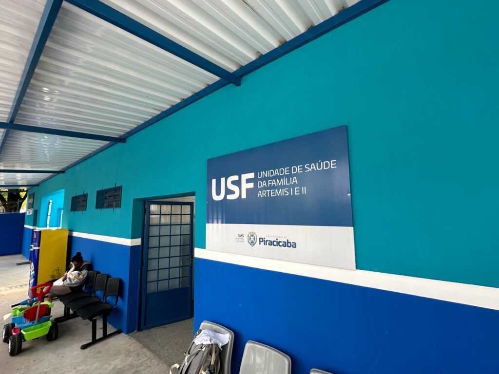 A imagem mostra a fachada de uma unidade de saúde familiar. A parede principal é pintada em um tom vibrante de azul turquesa, com uma faixa azul mais escura na parte inferior, separada por uma linha branca fina. Na parede azul turquesa, há uma placa grande e retangular com as seguintes informações em letras brancas: "USF UNIDADE DE SAÚDE DA FAMÍLIA ARTEMIS I E II". Abaixo disso, aparecem os logotipos da "SMS SECRETARIA DE SAÚDE" e da "PREFEITURA DE Piracicaba". À esquerda da placa, há uma porta de vidro com divisórias quadriculadas, que parece ser a entrada principal da unidade. Ao lado da porta, há duas janelas retangulares com grades. No lado esquerdo da imagem, é possível ver uma pessoa sentada em uma cadeira de plástico preta, virada para a esquerda. Ao lado da pessoa, há brinquedos coloridos, incluindo um carrinho de plástico verde e vermelho. Há também um dispensador de água amarelado e uma estrutura metálica azul cobrindo a área externa, com um teto de metal branco com ripas. Na parte inferior da imagem, em primeiro plano, há mais cadeiras de plástico cinza e uma mochila apoiada em uma delas. O chão parece ser de concreto. O ambiente geral sugere uma área de espera externa em um dia claro.