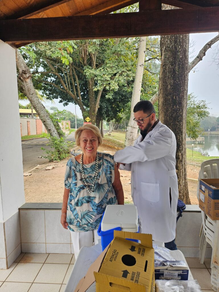 Nesta imagem, uma pessoa vestida com jaleco branco aplica uma injeção em outra que está de pé, em um ambiente ao ar livre. A cena ocorre sob uma estrutura de madeira com telhado, cercada por árvores e um caminho ao fundo. Sobre uma mesa próxima, há caixas e suprimentos médicos, sugerindo que se trata de uma ação de saúde, como uma campanha de vacinação. A atmosfera transmite cuidado e atenção à saúde pública em um contexto comunitário.