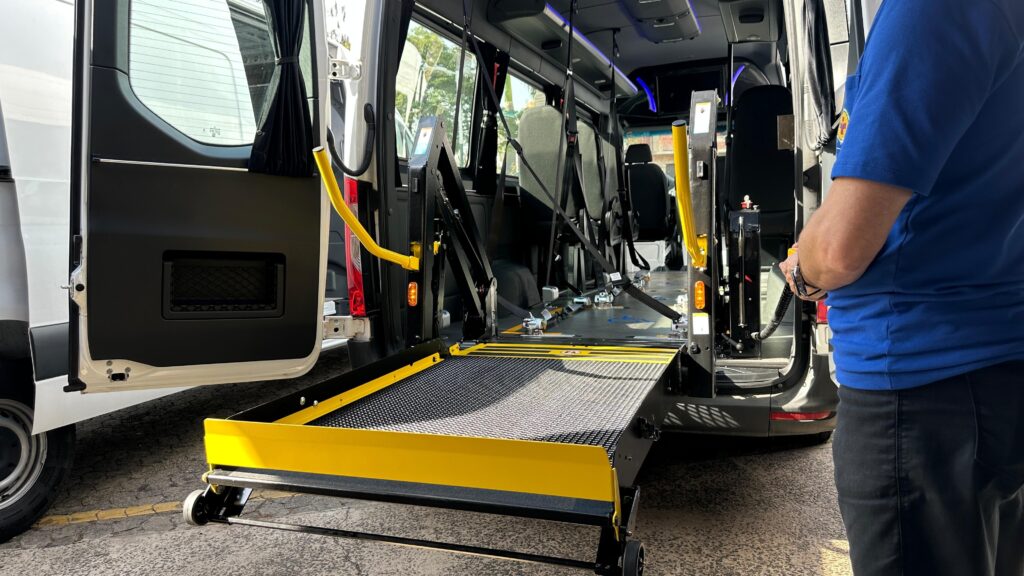 A imagem mostra uma van adaptada para acessibilidade, com uma plataforma elevatória para cadeiras de rodas em posição de uso. A plataforma, de cor amarela e preta, está estendida para fora do veículo, com detalhes em metal texturizado para aderência. O interior da van revela assentos com cintos de segurança e sistemas de fixação para cadeiras de rodas. Há também barras de apoio amarelas nas laterais e no interior do veículo, projetadas para auxiliar na mobilidade.