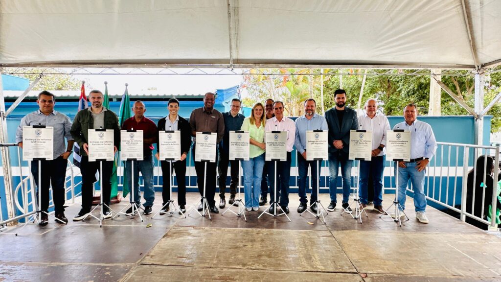A imagem mostra um grupo de pessoas em pé em um palco sob uma tenda. Todos estão vestidos de maneira formal ou semi-formal e seguram placas relacionadas a inauguração.Na frente do grupo, há vários suportes exibindo as placas de inauguração. O fundo apresenta uma parede azul e um pouco de vegetação, sugerindo um local ao ar livre ou semi-aberto.