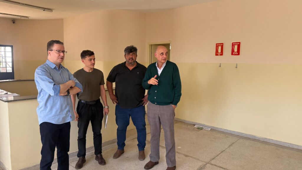 A imagem mostra um grupo de cinco homens em pé em frente a um carro da polícia científica estacionado na rua. O carro é branco com detalhes em preto e tem a inscrição "POLÍCIA CIENTÍFICA" no capô. Os homens parecem estar em uma área urbana, com um prédio ao fundo e uma parede de tijolos com grafites. O dia está ensolarado, com sombras projetadas no chão.O homem mais à esquerda está vestindo uma camisa azul clara e calças escuras. Ao seu lado, um homem com camisa branca e um distintivo pendurado no pescoço. O terceiro homem da esquerda usa uma camisa preta e calças azuis. O quarto homem está com uma camisa verde sobre uma camisa branca e calças marrons. O último homem, à direita, veste uma camiseta verde escura e calças pretas, segurando um objeto retangular na mão. Todos estão olhando para a câmera e parecem posar para a foto.