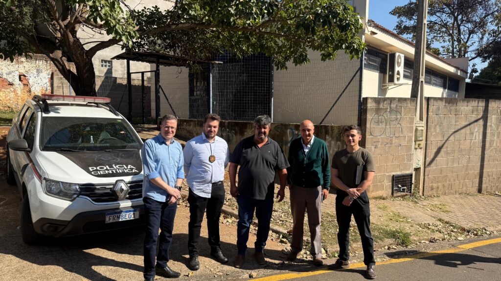 A imagem mostra um grupo de cinco homens em pé em frente a um carro da polícia científica estacionado na rua. O carro é branco com detalhes em preto e tem a inscrição "POLÍCIA CIENTÍFICA" no capô. Os homens parecem estar em uma área urbana, com um prédio ao fundo e uma parede de tijolos com grafites. O dia está ensolarado, com sombras projetadas no chão.O homem mais à esquerda está vestindo uma camisa azul clara e calças escuras. Ao seu lado, um homem com camisa branca e um distintivo pendurado no pescoço. O terceiro homem da esquerda usa uma camisa preta e calças azuis. O quarto homem está com uma camisa verde sobre uma camisa branca e calças marrons. O último homem, à direita, veste uma camiseta verde escura e calças pretas, segurando um objeto retangular na mão. Todos estão olhando para a câmera e parecem posar para a foto.
