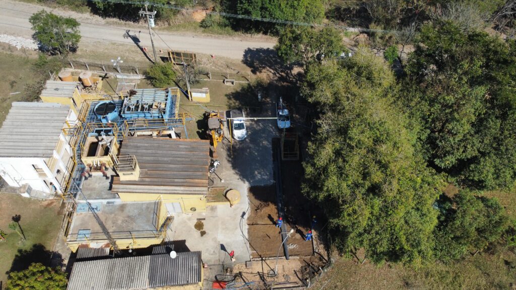 A imagem mostra uma vista aérea de uma estação de tratamento de água ou de uma instalação industrial semelhante. *Detalhes observados:* * *Edifícios:* Há vários edifícios com telhados de metal e concreto. Um edifício principal, mais branco, parece ser um escritório ou controle central. Outros edifícios e estruturas são pintados de amarelo e possuem telhados de metal com inclinação. * *Equipamentos:* Várias estruturas metálicas, incluindo plataformas, escadas, tubulações e tanques, estão visíveis. Há equipamentos de grande porte, como filtros ou decantadores, com componentes azuis e amarelos. * *Veículos:* Um trator de esteira (amarelo) e dois veículos utilitários brancos estão estacionados na área. * *Trabalhadores:* Várias pessoas vestindo uniformes azuis e capacetes estão trabalhando em uma área de terra batida, possivelmente realizando obras ou manutenção. * *Paisagem:* A instalação está cercada por vegetação exuberante, com muitas árvores de grande porte. Uma estrada de terra ou cascalho passa pela parte superior da imagem. * *Infraestrutura:* Postes de eletricidade e fiação são visíveis, indicando a presença de energia elétrica na instalação. A cena geral sugere uma operação industrial em andamento, com atividades de manutenção ou construção ocorrendo em uma área externa.