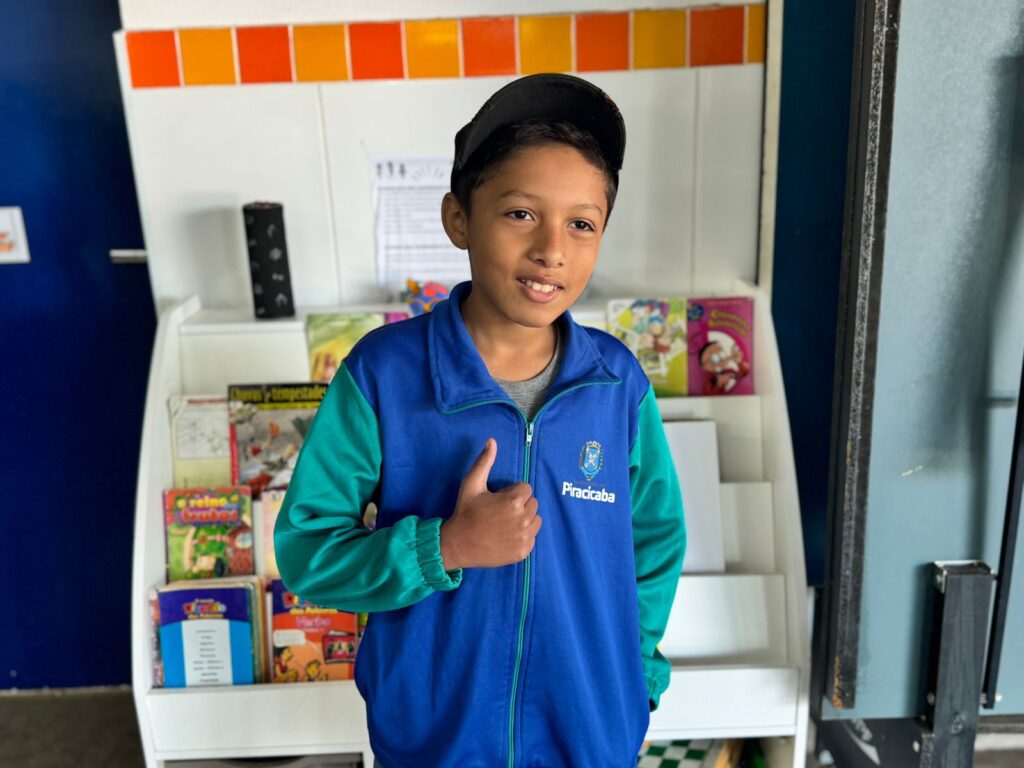 A imagem apresenta uma pessoa em pé diante de uma estante branca repleta de livros e revistas coloridas. Ela veste uma jaqueta azul e verde com a palavra “Piracicaba” estampada nas costas e faz um gesto de positivo com o polegar. O ambiente parece ser uma biblioteca ou espaço educativo, com uma parede decorada com azulejos laranja e amarelos na parte superior e uma porta azul à esquerda.