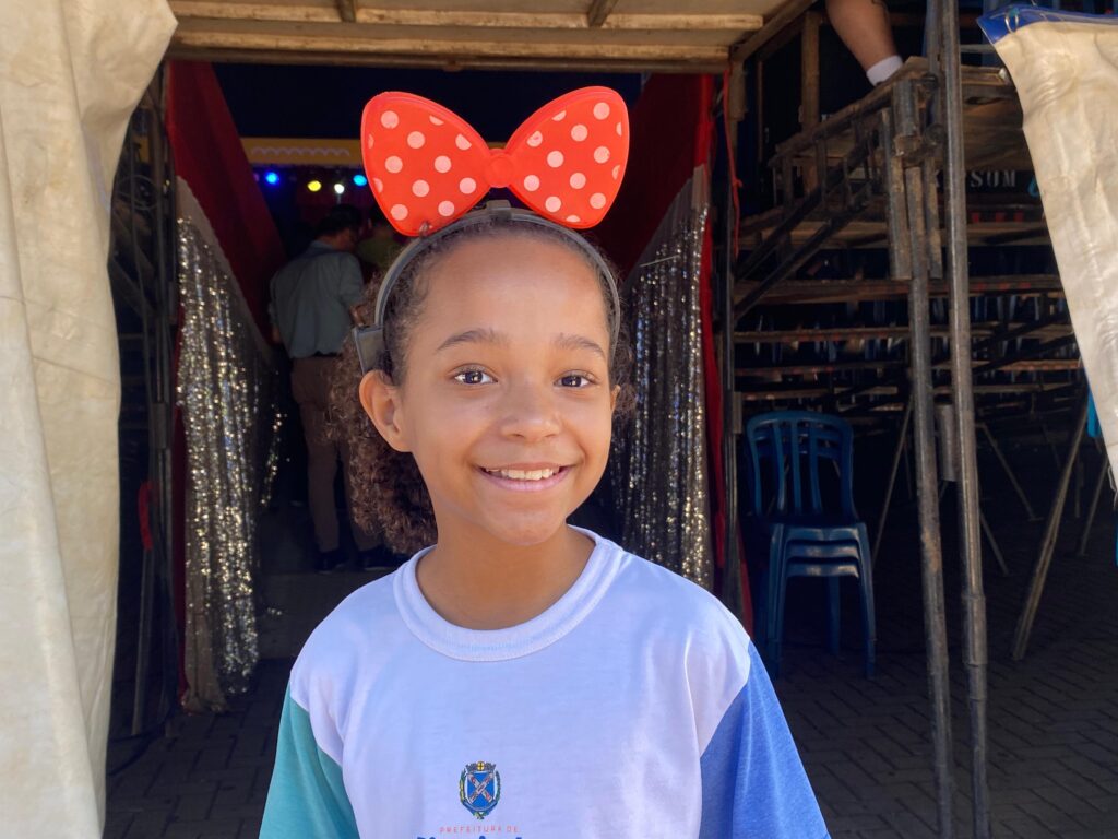 Menina negra de 8 anos sorri em frente à entrada do circo, usando uniforme escolar da Prefeitura de Piracicaba (camiseta branca com mangas azul e verde). Na cabeça, uma tiara vermelha com um laço grande de bolinhas brancas.