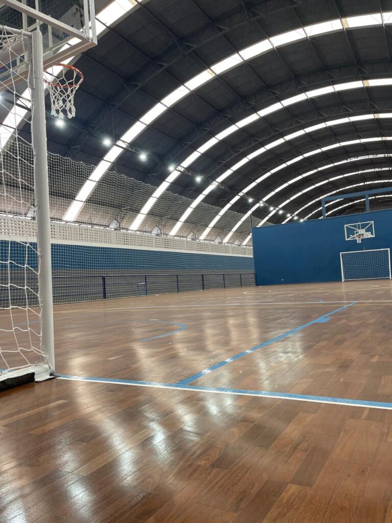 A imagem mostra o interior de uma quadra esportiva coberta, com piso de madeira polida e marcado com linhas azuis. O teto abobadado é sustentado por vigas metálicas escuras e iluminado por uma série de luminárias embutidas. À esquerda, parte de uma cesta de basquete com sua rede branca é visível. Ao fundo, a quadra se estende, com paredes azuis e redes de proteção, e um gol de futsal pode ser visto ao longe. A iluminação cria reflexos no piso brilhante, dando uma sensação de amplitude e limpeza ao espaço.