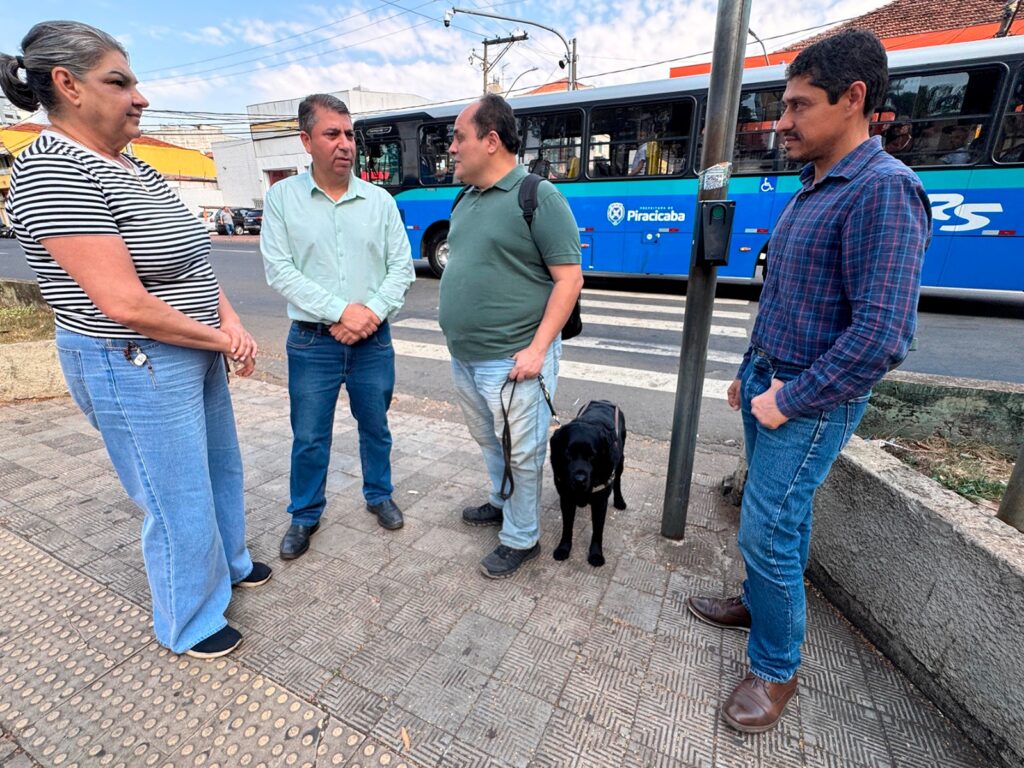 A imagem mostra quatro pessoas em pé em uma calçada, com um ônibus azul ao fundo. Uma mulher e três homens estão conversando, e um dos homens está segurando a coleira de um cachorro preto. Todos estão vestindo roupas casuais. O ônibus tem a palavra "Piracicaba" escrita em letras brancas. A calçada é feita de ladrilhos de concreto com um padrão geométrico. Há um poste com um semáforo e um poste de luz. O céu está parcialmente nublado.