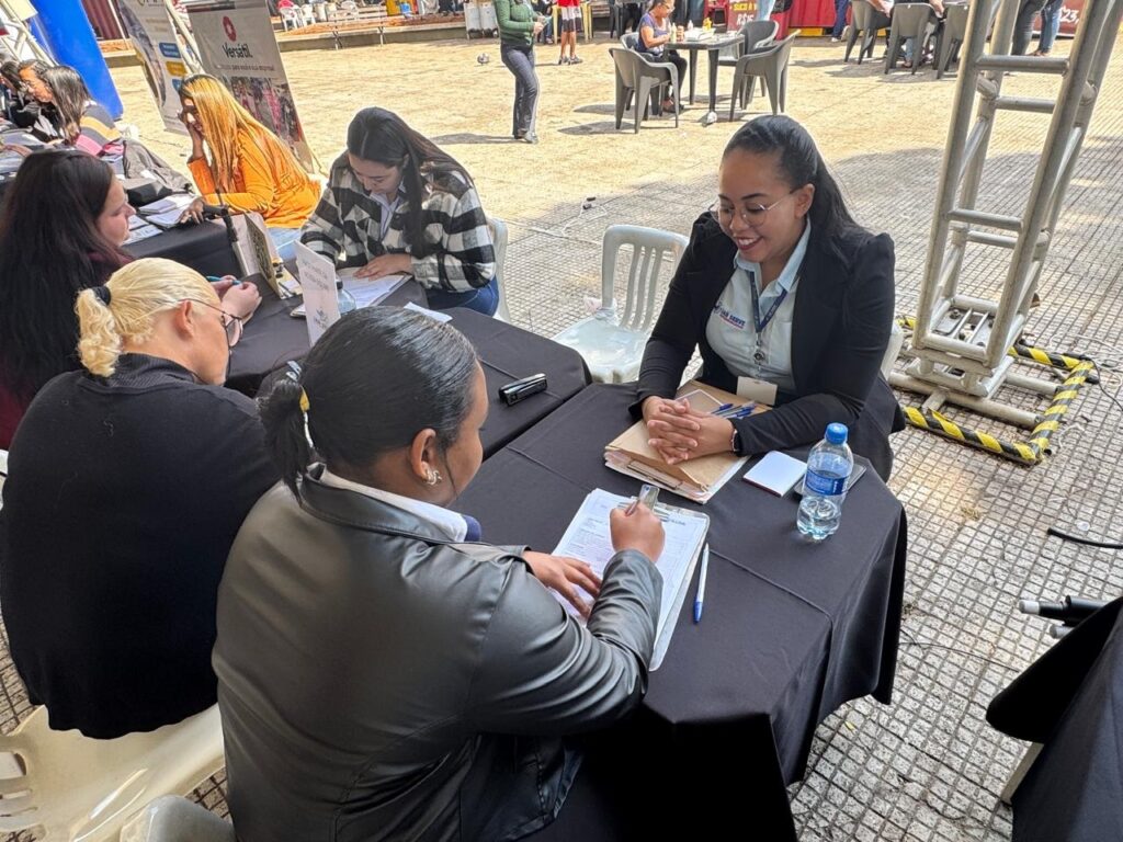 A imagem mostra um evento ao ar livre onde pessoas estão sentadas em mesas cobertas com toalhas pretas, participando de uma atividade. Há quatro mulheres visíveis sentadas à mesa principal. Uma mulher, vestindo um blazer preto e camisa azul clara com um crachá, sorri enquanto olha para baixo, com as mãos juntas sobre a mesa. Ela parece estar interagindo com alguém que está preenchendo um formulário. As outras três mulheres estão sentadas do outro lado da mesa, de costas para a câmera ou viradas de lado. Uma delas tem cabelo loiro preso em um coque e usa óculos. Outra tem cabelo preto preso em um rabo de cavalo e usa um casaco preto brilhante. A terceira mulher, mais à esquerda, tem cabelo preto longo e usa um suéter listrado. Ao fundo, outras pessoas estão em pé ou sentadas em mesas, e há banners e estruturas metálicas visíveis, sugerindo que se trata de um evento de recrutamento, feira de carreiras ou similar. O ambiente é iluminado pelo sol, indicando que é dia.