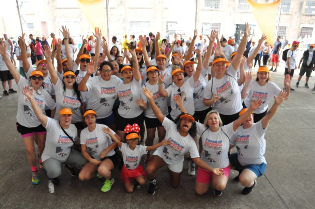 A imagem mostra um grande grupo de pessoas, a maioria mulheres, posando para uma foto. Eles estão todos vestindo camisetas brancas com um design semelhante e a maioria usa bonés ou viseiras laranja. Muitos deles estão acenando e parecem estar felizes e animados. O grupo parece estar participando de um evento, possivelmente uma caminhada ou corrida, como sugerido pelas camisetas que parecem ter um logotipo ou texto relacionado a um evento. O fundo mostra uma estrutura de construção, possivelmente um centro de convenções ou um local de evento ao ar livre, com algumas bandeiras alaranjadas visíveis ao fundo. Há uma mistura de idades no grupo, com adultos e algumas crianças presentes. A atmosfera geral é festiva e comunitária.