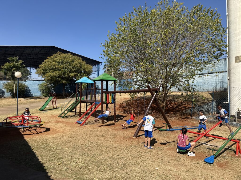 A imagem mostra um parquinho infantil ensolarado com várias crianças brincando. No centro, há uma estrutura de playground com dois escorregadores e um balanço. Uma menina está sentada em um dos balanços, enquanto um menino está em pé ao lado do outro balanço. Há também um carrossel à esquerda, onde uma criança está sentada. Ao fundo, um grande jogo de gangorra com várias crianças. O parquinho está cercado por grama e árvores, e o céu está claro e azul.