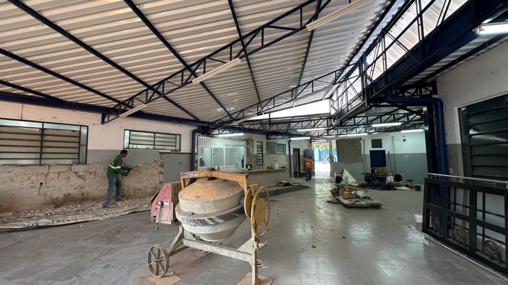 A imagem mostra o interior de um edifício em construção ou reforma. Há um trabalhador usando uma furadeira para demolir uma parede de tijolos. No centro da imagem, há uma betoneira. O teto é feito de telhas metálicas e sustentado por uma estrutura de metal azul. Há janelas com grades nas paredes. O chão é de cimento. Há materiais de construção espalhados pelo local, como sacos de cimento, caixas e ferramentas.