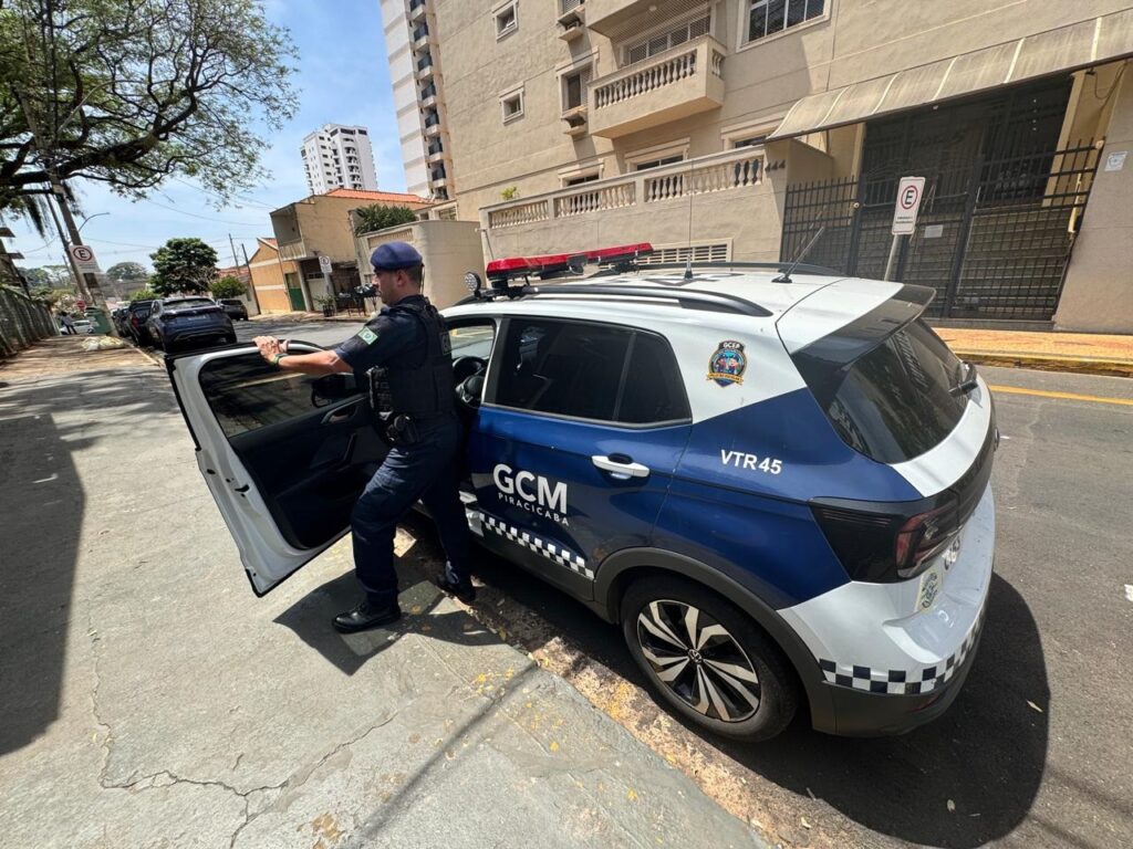 A imagem mostra um policial uniformizado saindo de um carro oficial da Guarda Civil Municipal (GCM) de Piracicaba. O policial está com a porta do motorista aberta e com a mão apoiada no teto do carro, enquanto seu pé esquerdo está no chão. Ele veste um colete tático sobre o uniforme escuro e usa um boné azul. O carro é um SUV branco com detalhes em azul, com as inscrições "GCM PIRACICABA" e "VTR 45" visíveis. Na traseira do veículo, há um padrão xadrez preto e branco, característico de veículos de segurança. O cenário é urbano, com um prédio residencial ao fundo, árvores e outras árvores e carros estacionados na rua. O dia parece claro e ensolarado.