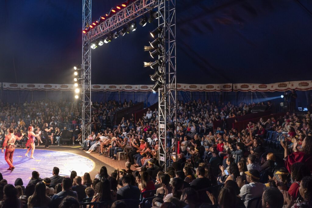 Público lota arquibancadas e cadeiras em uma grande lona de circo iluminada.
