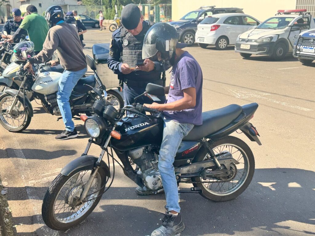 A polícia aborda um motociclista em uma rua ensolarada. O policial, vestindo uniforme azul escuro e colete tático, está de pé e parece estar verificando os documentos do motociclista, que está sentado em sua moto preta. O motociclista usa um capacete preto, camiseta roxa e calça jeans clara. Outras motocicletas e carros estão estacionados ao fundo, e algumas pessoas podem ser vistas andando na calçada. O sol projeta sombras nítidas no asfalto.