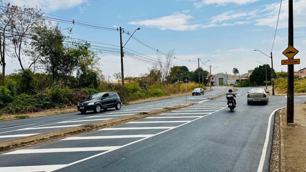 A imagem mostra uma rua com tráfego em um dia ensolarado. Há vários carros e uma motocicleta na estrada. A estrada é pavimentada e tem faixas brancas pintadas. Há também uma faixa de pedestres na estrada. A vegetação é visível em ambos os lados da estrada, com árvores e arbustos. Vários postes de eletricidade com fios correm ao longo da estrada. Um sinal de trânsito amarelo com um símbolo de lombada e "A 150m" é visível em um poste. O céu está azul com algumas nuvens brancas.