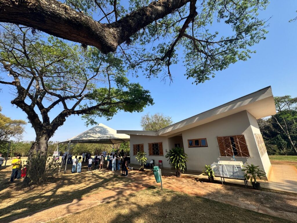 Um prédio moderno de um andar com telhado inclinado está cercado por árvores grandes e grama verde. Ao lado do prédio, há uma tenda onde várias pessoas estão reunidas, indicando que está acontecendo um evento social ou público. O prédio tem janelas com molduras de madeira e vasos de plantas decorando a entrada. O céu está limpo e azul, sugerindo um dia ensolarado.