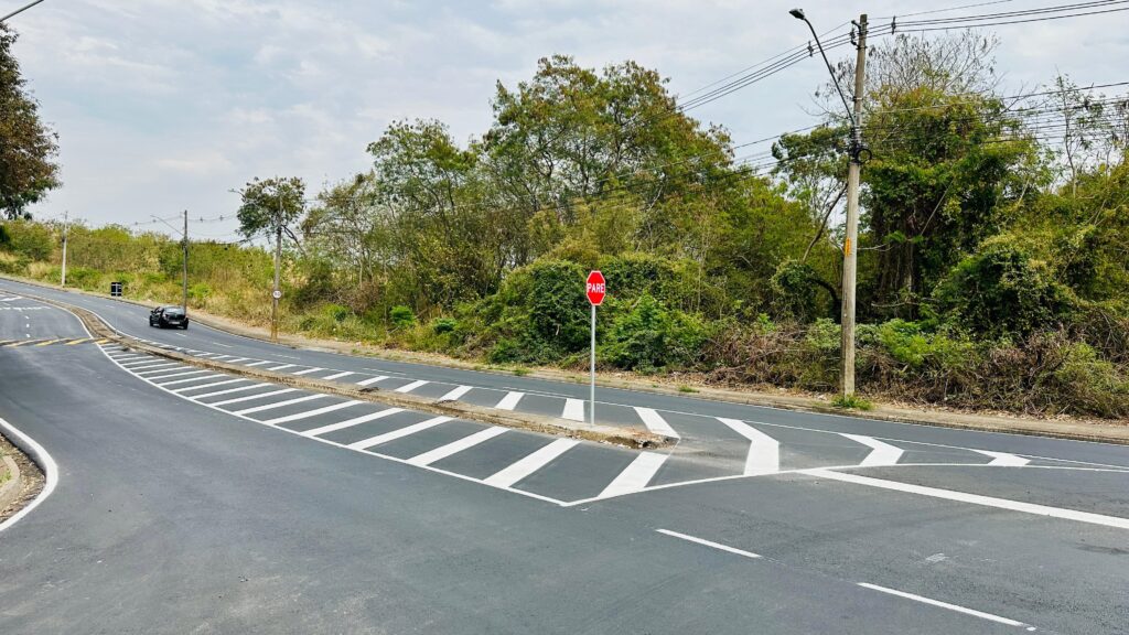 A imagem mostra uma estrada com várias faixas de rolamento, com uma curva suave à esquerda e uma interseção à direita. Há um sinal de "PARE" vermelho e octogonal posicionado em uma ilha de tráfego no lado direito da estrada. A estrada é pavimentada com asfalto e marcada com linhas brancas, incluindo linhas de desaceleração ou de transição para as faixas da direita. Ao fundo, há uma vegetação densa com árvores e arbustos, com alguns fios elétricos e um poste de iluminação pública visíveis no lado direito. O céu está nublado, com algumas nuvens dispersas. Um carro preto está circulando na faixa da esquerda, afastando-se da câmera. No lado esquerdo da estrada, há uma área com vegetação mais baixa e um pequeno sinal de trânsito. A ilha de tráfego onde está o sinal de "PARE" é elevada e cercada por concreto. Há também algumas linhas brancas em zigue-zague na estrada, indicando uma área de perigo ou de mudança de pista.