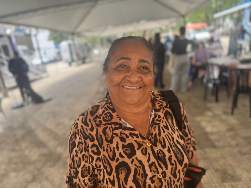 A mulher sorri abertamente para a câmera, com a pele enrugada e olhos brilhantes. Ela usa uma blusa de oncinha com botões dourados e um colar fino. O fundo está desfocado, mas mostra uma tenda branca, pessoas em plano de fundo e alguns veículos. Parece ser um local ao ar livre, talvez um evento ou mercado.