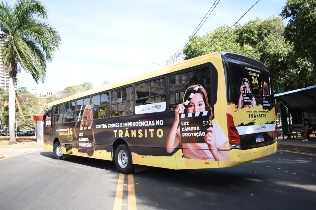 A imagem mostra a lateral traseira de um ônibus amarelo decorado com uma campanha de segurança no trânsito. O ônibus exibe a mensagem "CONTRA CRIMES E IMPRUDÊNCIAS NO TRÂNSITO" em letras grandes e amarelas sobre um fundo preto. Há também imagens de uma jovem segurando uma claquete e a inscrição "LUZ CÂMERA PROTEÇÃO", indicando o uso de câmeras de vigilância para aumentar a segurança. A frase "CIDADE MONITORADA 24 HRS" reforça a ideia de vigilância contínua. Ao fundo, vê-se uma parada de ônibus com pessoas sentadas e árvores frondosas, sugerindo um ambiente urbano. O céu está claro e azul, com algumas nuvens.