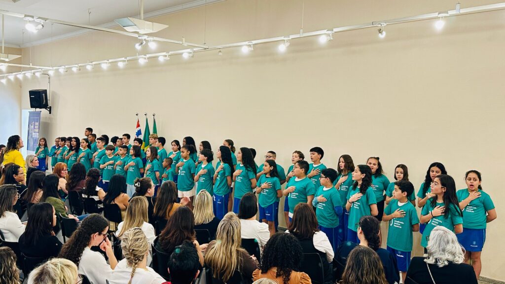 A imagem mostra um grupo de crianças e adolescentes em pé, em frente a uma plateia sentada. Eles estão vestindo camisetas verde-água com uma inscrição em português que parece ser "Academia Jovens Músicos". A maioria deles está com a mão direita sobre o peito, em um gesto que pode indicar respeito, como cantar o hino nacional ou expressar patriotismo. Ao fundo, há bandeiras do Brasil e de outros países, sugerindo um evento ou apresentação cultural. As crianças e adolescentes parecem estar cantando ou se apresentando para a audiência, que é composta principalmente por adultos. A iluminação do ambiente é feita por um sistema de spots no teto, e a parede ao fundo é clara, criando um ambiente formal para a ocasião.