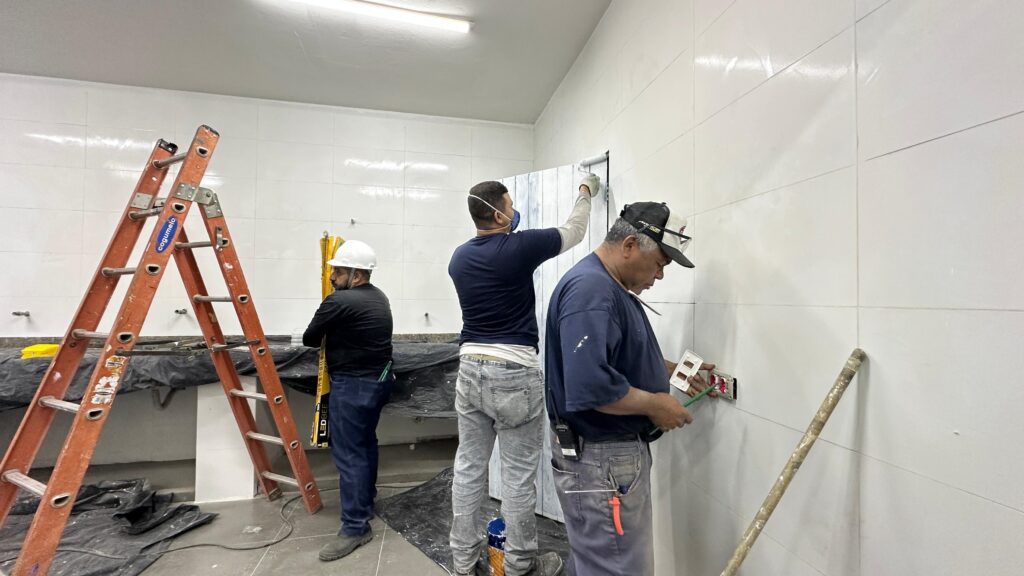 A imagem mostra três homens trabalhando em uma sala com paredes de azulejo branco. À esquerda, um homem com capacete branco e camisa escura está em frente a uma escada laranja. A escada está encostada em uma parede e há lonas pretas cobrindo o chão e o balcão abaixo. No centro, um homem com máscara de proteção e camiseta azul escura está pintando uma porta de madeira com listras verticais brancas e cinzas. Ele está usando luvas e tem uma lata de tinta no chão. À direita, um homem com boné e camiseta azul escura está mexendo em uma tomada na parede. Ele está segurando uma ferramenta verde e parece estar instalando ou consertando a tomada. Há um cano encostado na parede ao lado dele.