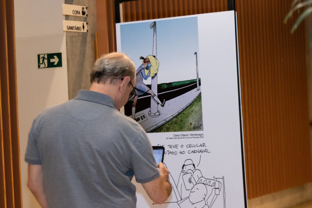 A imagem mostra um homem de costas, usando uma camisa polo cinza, olhando para uma exposição de cartuns. Ele está segurando um celular na mão direita, como se estivesse tirando uma foto ou filmando a obra. No fundo, há uma parede branca com placas de sinalização. Uma placa indica "COPA" com um ícone de futebol, e outra indica "SANITÁRIO" com um ícone de uma pessoa. Há também uma placa de saída de emergência verde com um homem correndo em direção a uma seta. A exposição em si é um grande cartum colorido, com a assinatura "Darko Drljevic | Montenegro" e a informação "50° Salão Internacional de Humor de Piracicaba 2023". O cartum retrata uma pessoa com uma máscara, usando um macacão azul e branco, com um "AIR BAG" escrito em seu joelho, correndo e se agarrando a um poste. Abaixo deste cartum principal, há um desenho em preto e branco de duas pessoas deitadas, uma delas segurando um celular e dizendo "FITEVE O CELULAR UTADO NO CARNAVAL".
