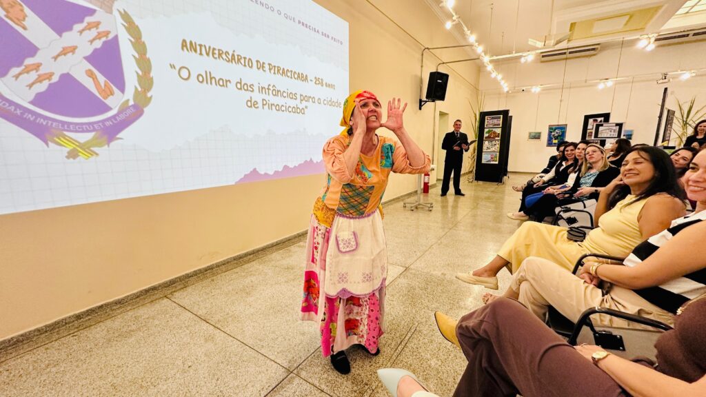 A imagem mostra uma sala onde um evento está acontecendo. No centro, uma mulher com uma roupa colorida e um lenço na cabeça está gesticulando com as mãos levantadas, como se estivesse falando ou cantando. Atrás dela, um homem de terno está em pé, possivelmente apresentando algo. Ao fundo, há um telão com o texto "ANIVERSÁRIO DE PIRACICABA - 258 anos" e "O olhar das infâncias para a cidade de Piracicaba", além de um brasão com peixes. Uma audiência está sentada em cadeiras, voltada para a apresentação, com algumas pessoas sorrindo. O ambiente parece ser um local de eventos ou uma galeria, com quadros expostos nas paredes e iluminação de teto.
