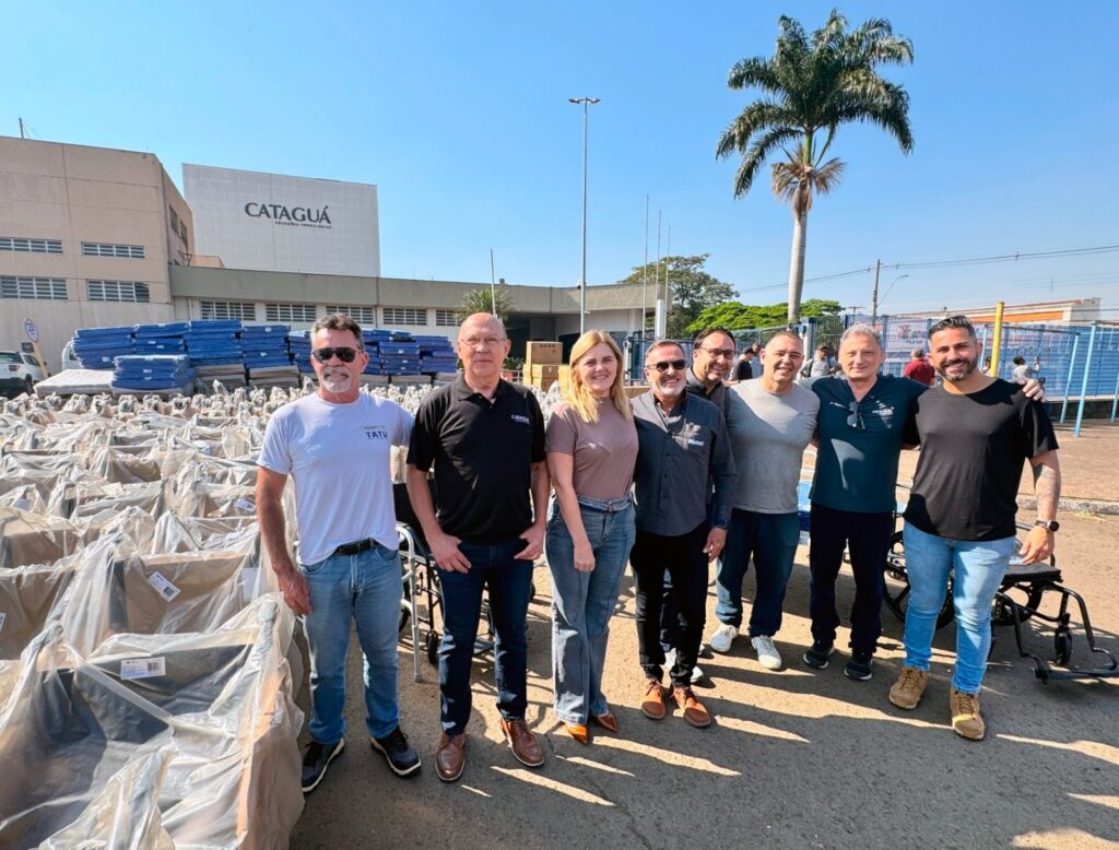 Na imagem, um grupo de oito pessoas posa em frente a um edifício com a inscrição "CATAGUÁ". Há também uma grande quantidade de materiais embalados, possivelmente para doação ou distribuição, atrás do grupo. As pessoas estão em pé, sorrindo para a câmera, e a maioria delas está vestida casualmente. O céu está claro e azul, com uma palmeira proeminente ao fundo, sugerindo um dia ensolarado.