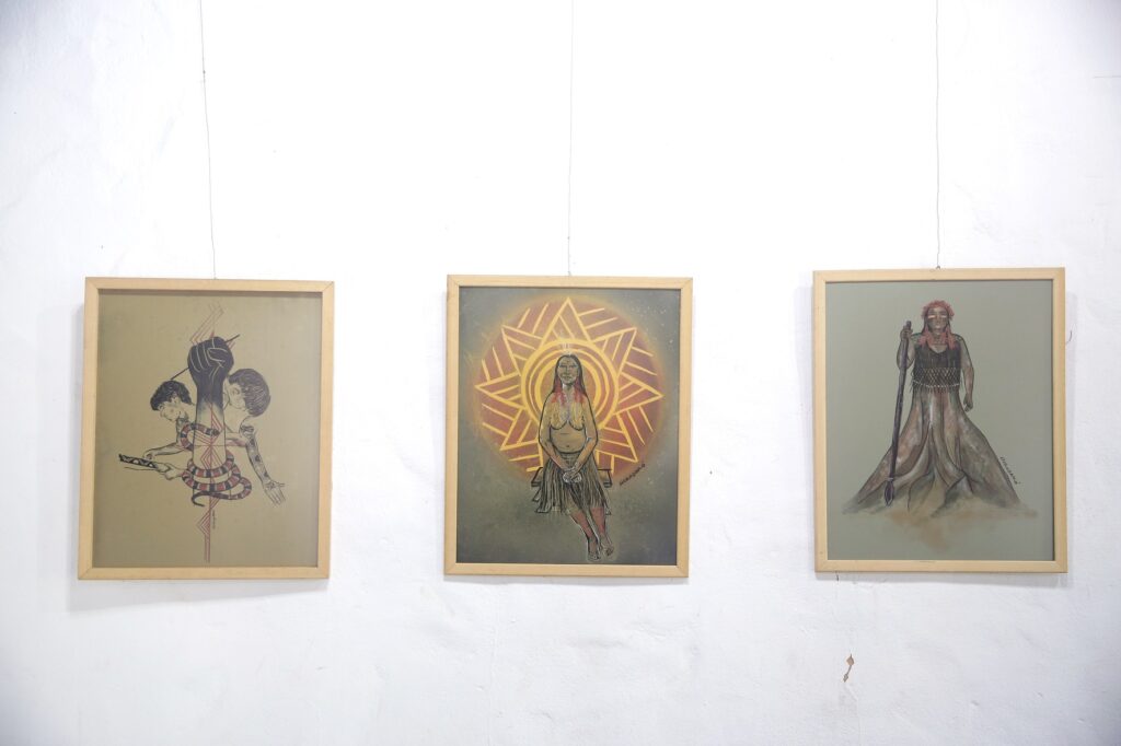 A imagem exibe três quadros pendurados em uma parede branca e texturizada. Cada quadro é emoldurado em madeira clara e contém uma obra de arte distinta. O quadro da esquerda apresenta um desenho em tons terrosos com figuras estilizadas. No centro, um punho cerrado emerge, com linhas vermelhas e pretas irradiando dele. Abaixo do punho, duas cabeças humanas estão representadas, uma de perfil e outra de frente, conectadas por um corpo envolto em uma serpente. Uma das figuras parece estar manipulando um objeto retangular com linhas em zigue-zague. O quadro central é uma pintura vibrante com uma figura feminina em destaque. Ela está sentada, com os olhos fechados e um leve sorriso, adornada com colares e com os cabelos longos e escuros. Atrás dela, um grande sol estilizado em tons de laranja e amarelo cria um ambiente radiante. A figura tem a pele clara e veste uma saia longa e escura. Há uma assinatura na parte inferior direita da pintura.