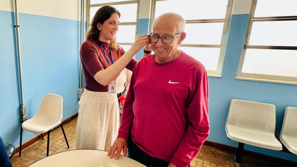 Uma mulher de cabelos castanhos e um homem mais velho com óculos estão em uma sala com paredes azuis claras. A mulher, que usa uma blusa de manga comprida em tom de vinho e uma saia bege, está ajustando os óculos do homem. O homem veste uma camiseta de manga comprida vermelha com o logo da Nike. Ao fundo, há janelas e cadeiras brancas simples.